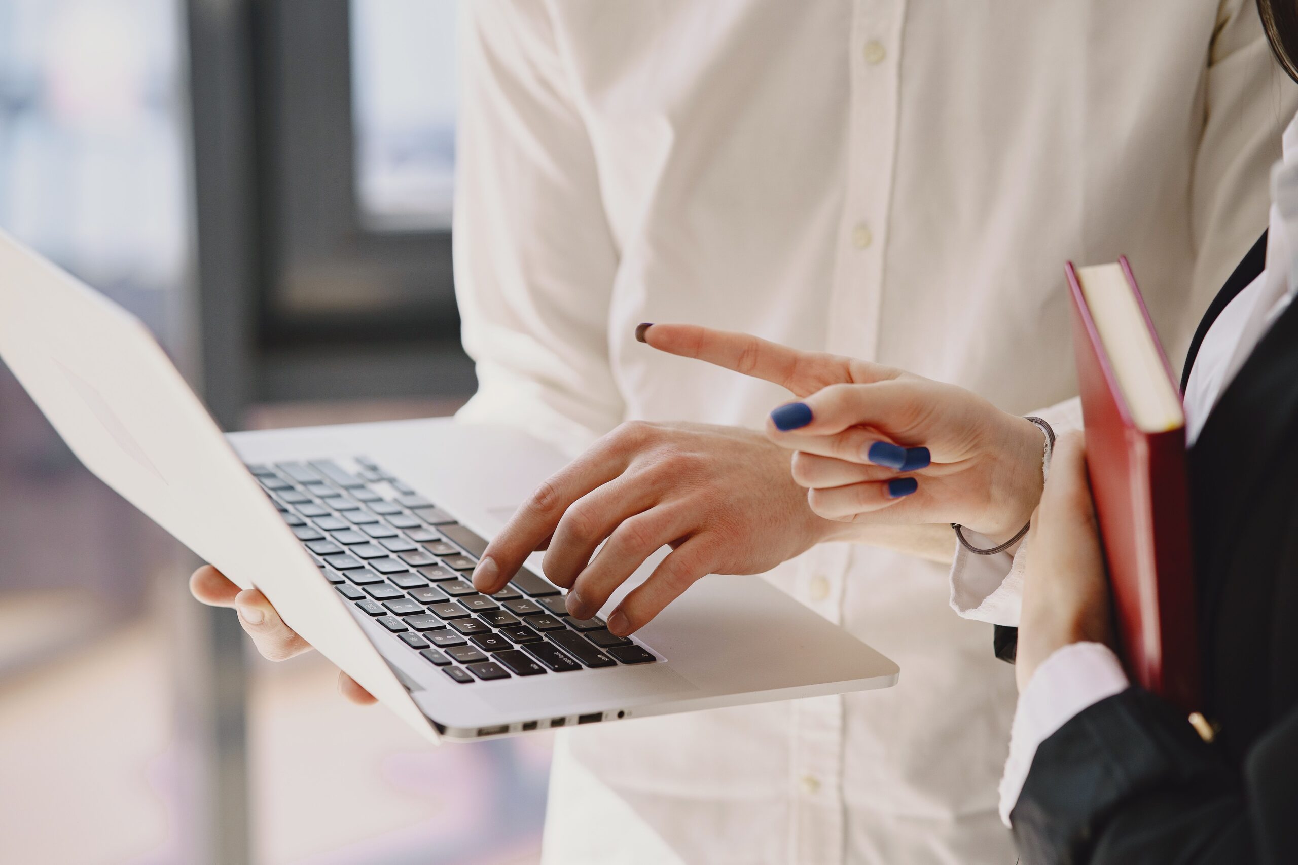 Crop well dressed anonymous male typing on netbook while standing near female partner showing with finger and taking copybook at work in daylight